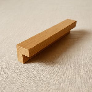 Könnte beinhalten: Ein hellbrauner Möbelgriff aus Holz mit einem linearen, rechteckigen Profil und einer glatten Oberfläche mit natürlicher Maserung. Das Beschlagteil hat ein L-förmiges Design mit einer flachen Oberseite und einer vertikalen Montagekante aus Massivholz.