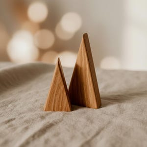 Może przedstawiać: Dwa drewniane przedmioty dekoracyjne w kształcie trójkąta. Obiekty wykonane są z jasnobrązowego drewna i mają naturalne usłojenie. Umieszczone są na tkaninie w neutralnym kolorze, z rozmytym tłem z miękkiego, ciepłego światła.