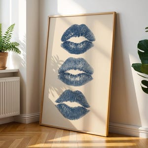 May include: A framed print with three blue lip imprints on a cream background. The artwork is in a light wooden frame, leaning against a white wall. The room has a wooden floor and a potted plant. The print is a stylish home decor item.