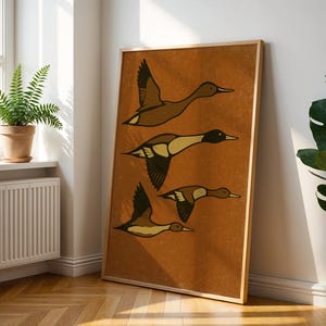 Pu&ograve; includere: Una stampa artistica incorniciata con un'illustrazione stilizzata di tre anatre in volo. Le anatre sono raffigurate in tonalit&agrave; di marrone, beige e nero su uno sfondo caldo marrone-arancio. L'opera d'arte &egrave; esposta in una stanza con luce naturale.
