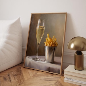 May include: A framed print depicts a champagne flute and a metal cup filled with french fries on a white tablecloth. The painting has a warm color palette and is set against a neutral background. A gold lamp and books are in the background.