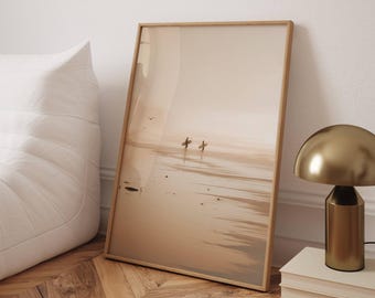 Op de afbeelding: Een ingelijste prent met een strandscène met twee figuren die surfplanken dragen. Het kunstwerk heeft een warm, neutraal kleurenpalet. Een goudkleurige lamp en een stapel boeken staan op een wit oppervlak.
