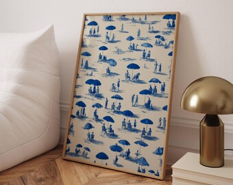 May include: A framed print featuring a blue and white beach scene with people, umbrellas, and beach chairs. The artwork has a vintage, toile-inspired design. The frame is a light wood color, and the print is leaning against a white wall.