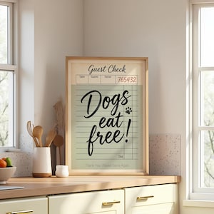 May include: A framed print with the words "Dogs eat free!" in a cursive font. The print resembles a guest check, with lines and a paw print. The frame is a light wood color, and the background is a kitchen setting.