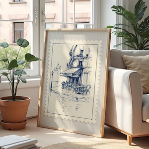 May include: A framed print depicting a blue and white sketch of a European cafe. The artwork is in a light wood frame and is propped against a wall near a window. A potted plant and a sofa are also visible.