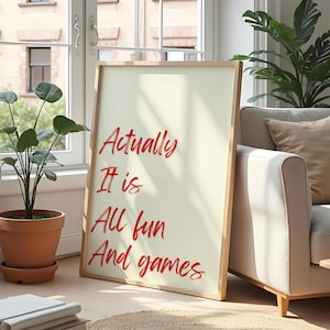 Pode incluir: Uma impressão emoldurada com as palavras "Actually It is All fun And games" em vermelho. A moldura é de madeira clara e o fundo é creme. A impressão está encostada em um sofá branco em uma sala bem iluminada.