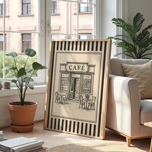 May include: Framed black and white illustration of a cafe with tables and chairs outside. The word "CAF&Eacute;" is above the entrance. The frame has a light wood color and black vertical stripes.