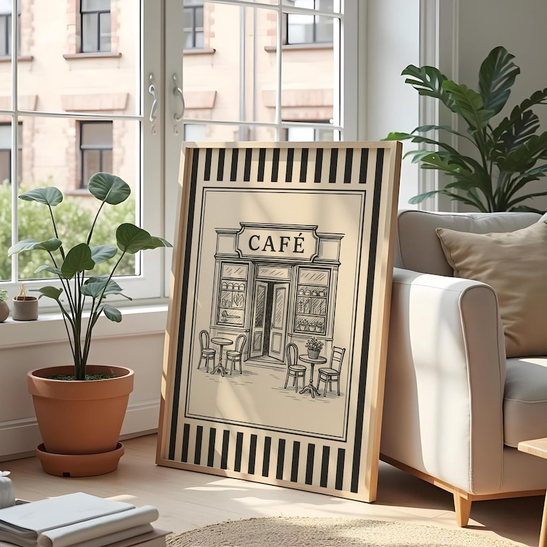 May include: Framed black and white illustration of a cafe with tables and chairs outside. The word "CAF&Eacute;" is above the entrance. The frame has a light wood color and black vertical stripes.