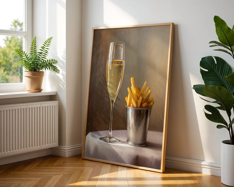 May include: A framed painting depicts a champagne flute filled with bubbly liquid and a metal cup overflowing with golden french fries. The artwork is leaning against a white wall, with a fern in a pot visible in the background.