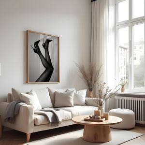 May include: A framed black and white photograph of legs in sheer stockings and high heels. The artwork hangs above a white sofa in a living room setting. The room has a neutral color palette.