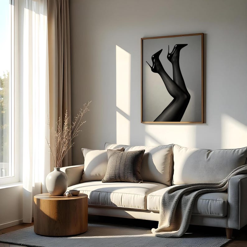 May include: A framed black and white photograph of legs in sheer black stockings and high heels hangs on a wall. The image is in a room with a beige sofa, a wooden side table, and a window with sheer curtains.