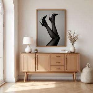 May include: A framed black and white art print featuring a pair of legs in black sheer stockings and high heels. The artwork is displayed above a wooden cabinet with a lamp, plant, and decorative items.