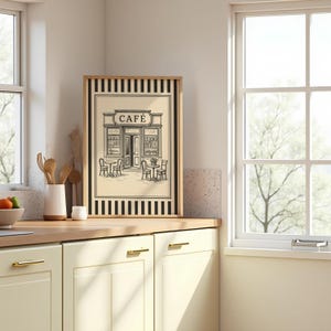 May include: Framed black and white print of a cafe scene. The artwork features a cafe facade with tables and chairs outside. The word "CAF&Eacute;" is at the top of the print. The frame is wood-toned with black and white striped borders.