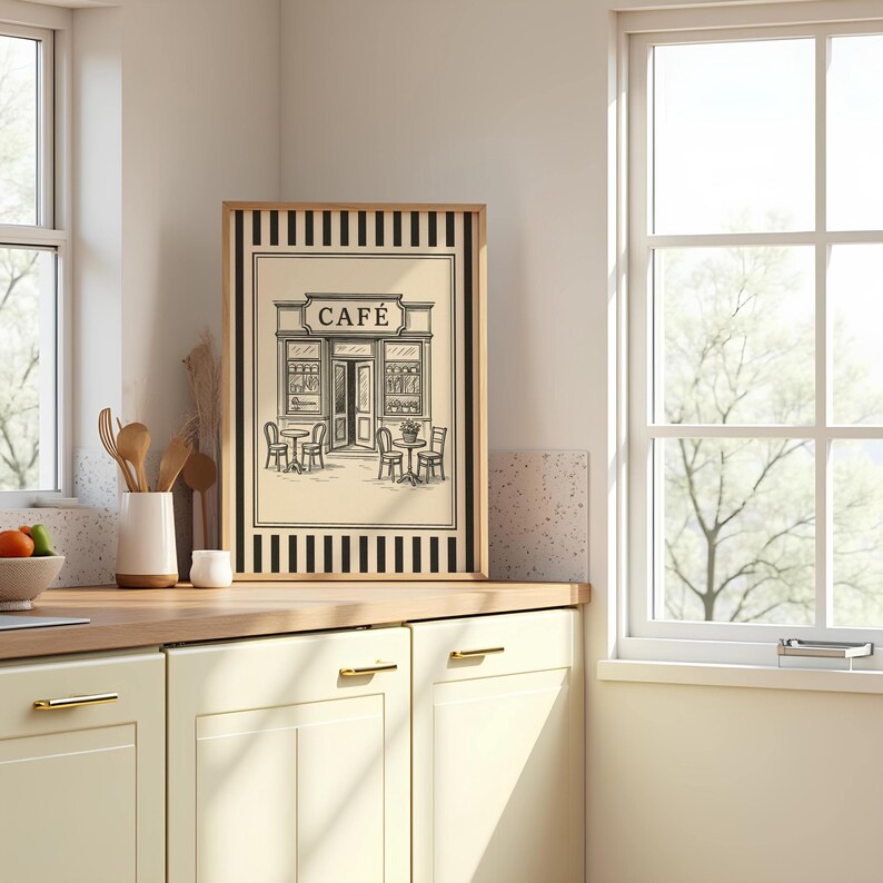 May include: Framed black and white print of a cafe scene. The artwork features a cafe facade with tables and chairs outside. The word "CAF&Eacute;" is at the top of the print. The frame is wood-toned with black and white striped borders.