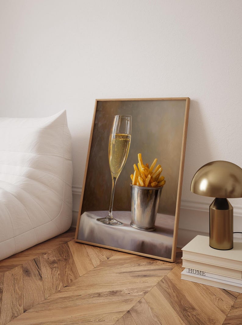 May include: A framed print depicts a champagne flute and a metal cup filled with french fries on a white tablecloth. The painting has a warm color palette and is set against a neutral background. A gold lamp and books are in the background.