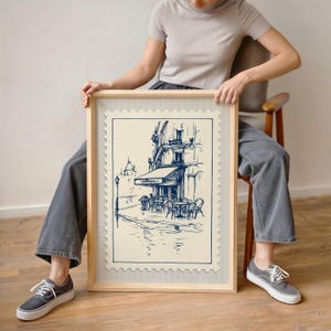 May include: A framed print showing a blue and white sketch of a European cafe. The artwork, in a postage stamp style, depicts a street scene with tables, chairs, and a building with an awning. The frame is made of light-coloured wood.