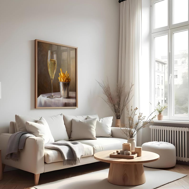 May include: A framed artwork depicts a champagne flute and a metal cup filled with french fries on a white tablecloth. The artwork hangs above a white sofa with pillows and a gray blanket. A wooden coffee table and a white ottoman are in front of the sofa.