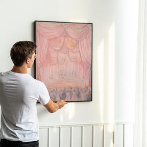 May include: A person hanging a framed painting on a white wall. The painting depicts a stage with pink and white curtains and a group of people in the background.