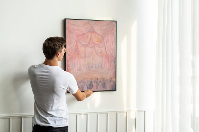 May include: A person hanging a framed painting on a white wall. The painting depicts a stage with pink and white curtains and a group of people in the background.