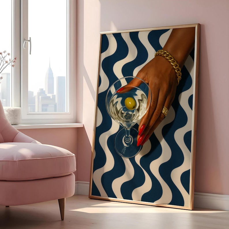 Peut inclure: Une impression d'art encadr&eacute;e repr&eacute;sentant une main tenant un verre &agrave; martini avec une olive. La main, orn&eacute;e de bijoux dor&eacute;s et de vernis &agrave; ongles rouge, est pos&eacute;e sur un motif ondul&eacute; bleu et blanc. L'&oelig;uvre est dans une pi&egrave;ce avec un mur rose et une vue sur la ville.