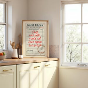 May include: A framed guest check print with the text "Chef was cute, would eat here again" in red. The print features a coffee stain and star ratings. The frame is light brown, set in a kitchen environment.