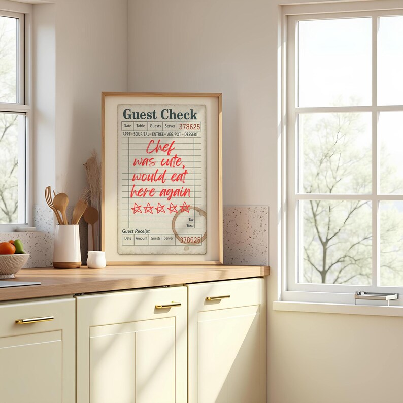 May include: A framed guest check print with the text "Chef was cute, would eat here again" in red. The print features a coffee stain and star ratings. The frame is light brown, set in a kitchen environment.