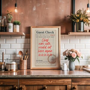 May include: A framed "Guest Check" print with red handwritten text, including "Chef was cute, would eat here again." The print is displayed on a wooden countertop with kitchen decor.