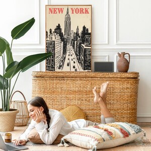 May include: A woman is sitting on a woven rug with a laptop computer, a cup of coffee, and a throw pillow.  She is looking at the laptop screen.  Behind her is a wicker chest with a framed print of a New York City skyline that says "NEW YORK" in red letters.