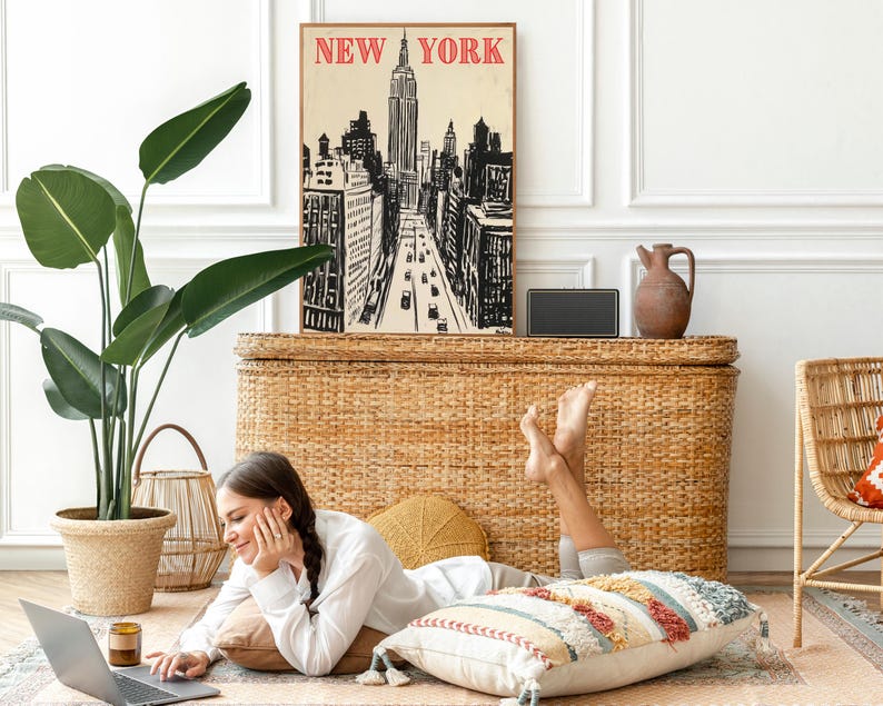 May include: A woman is sitting on a woven rug with a laptop computer, a cup of coffee, and a throw pillow.  She is looking at the laptop screen.  Behind her is a wicker chest with a framed print of a New York City skyline that says "NEW YORK" in red letters.