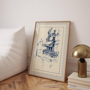 May include: A framed print showing a blue ink sketch of a Parisian cafe. The artwork, set against a cream background, depicts a street scene with tables, chairs, and a building with an awning. The frame is light wood, and the print is displayed on a wooden floor.
