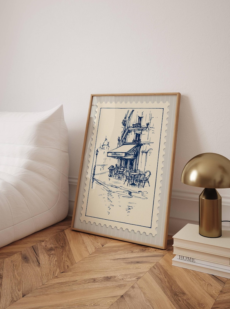 May include: A framed print showing a blue ink sketch of a Parisian cafe. The artwork, set against a cream background, depicts a street scene with tables, chairs, and a building with an awning. The frame is light wood, and the print is displayed on a wooden floor.
