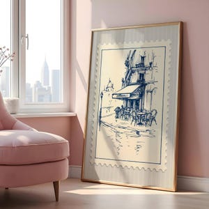 May include: A framed print showing a blue and white sketch of a cafe scene. The artwork, resembling a postage stamp, depicts a building with an awning, tables, and chairs. The frame is a light brown colour, and the background is a light beige.