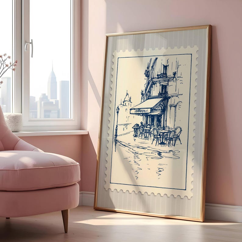 May include: A framed print showing a blue and white sketch of a cafe scene. The artwork, resembling a postage stamp, depicts a building with an awning, tables, and chairs. The frame is a light brown colour, and the background is a light beige.