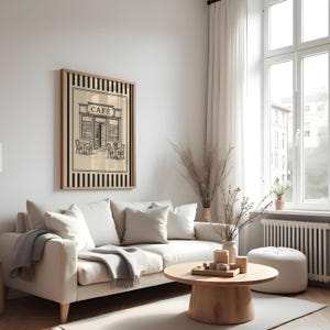 May include: A framed print of a cafe scene in black and white, with the word "CAFE" above the entrance. The print is in a wooden frame with black and beige stripes. The print hangs above a white sofa with pillows and a gray throw blanket.
