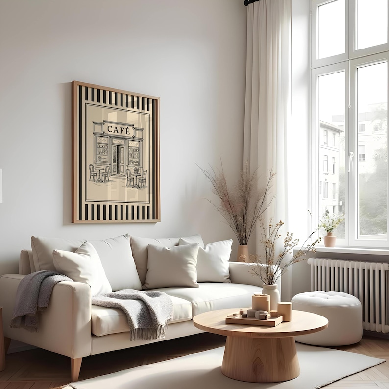 May include: A framed print of a cafe scene in black and white, with the word "CAFE" above the entrance. The print is in a wooden frame with black and beige stripes. The print hangs above a white sofa with pillows and a gray throw blanket.