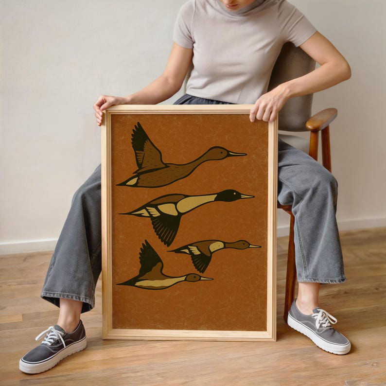 Pu&ograve; includere: Una stampa artistica incorniciata con un'illustrazione marrone e beige di tre anatre in volo. Le anatre hanno ali dettagliate e colli lunghi su uno sfondo strutturato marrone-arancio. L'opera &egrave; racchiusa in una cornice di legno naturale.