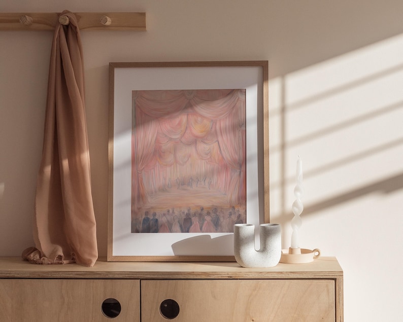 May include: A framed painting of a stage with pink and white curtains and a crowd of people in the background. The painting is hanging on a light wood wall with a brown scarf hanging on hooks above it. A white ceramic vase and a white candle are on the shelf below the painting.