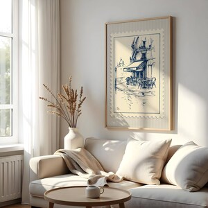 May include: A framed print depicting a cafe scene in a stamp design. The artwork shows a blue and white sketch of a European cafe with outdoor seating. The print is displayed above a cream sofa with cushions, a round wooden coffee table, and a white vase with dried plants.