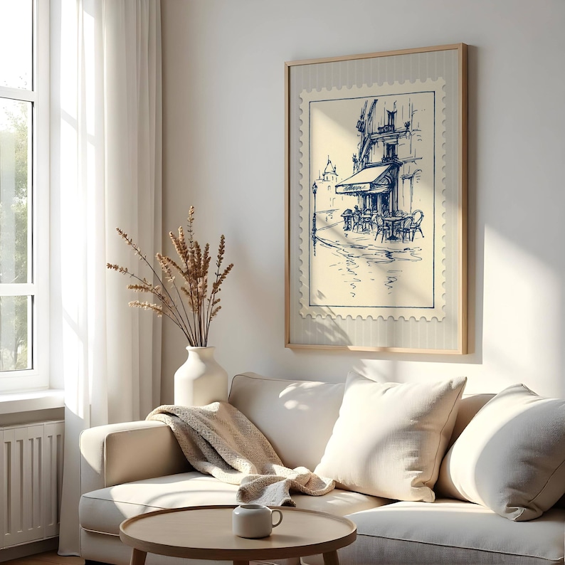 May include: A framed print depicting a cafe scene in a stamp design. The artwork shows a blue and white sketch of a European cafe with outdoor seating. The print is displayed above a cream sofa with cushions, a round wooden coffee table, and a white vase with dried plants.