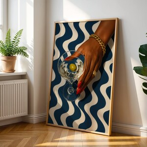 Peut inclure: Impression d'art encadr&eacute;e repr&eacute;sentant une main tenant un verre &agrave; martini avec une olive. La main est orn&eacute;e de bijoux dor&eacute;s et de vernis &agrave; ongles rouge. Le fond pr&eacute;sente un motif ondul&eacute; bleu et blanc.