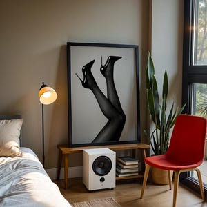 May include: A framed black and white print of legs in black stockings and high heels. The print is displayed above a small wooden bench with a white speaker and a stack of books. A red chair and a floor lamp are also in the room.