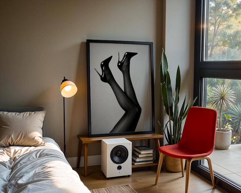 May include: A framed black and white print of legs in black stockings and high heels. The print is displayed above a small wooden bench with a white speaker and a stack of books. A red chair and a floor lamp are also in the room.