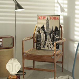May include: A black and white illustration of the New York City skyline with the Empire State Building in the center. The illustration is framed and hanging on a wall above a wooden chair. The chair is in a room with a wooden floor and a white wall.