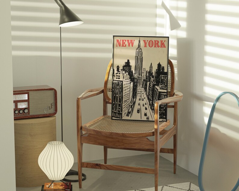 May include: A black and white illustration of the New York City skyline with the Empire State Building in the center. The illustration is framed and hanging on a wall above a wooden chair. The chair is in a room with a wooden floor and a white wall.