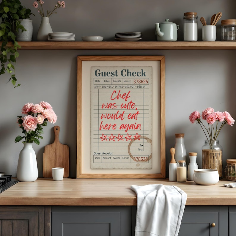 May include: A framed print of a restaurant guest check with the text "Chef was cute, would eat here again" in red. The print is displayed on a wooden countertop with kitchen accessories and pink flowers.