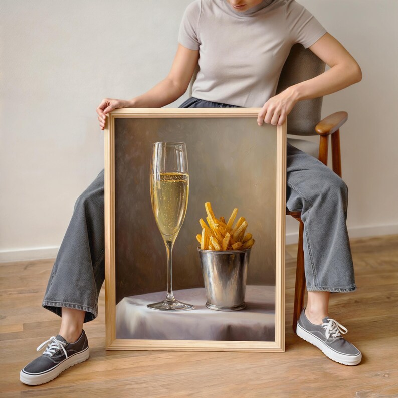 May include: A framed still life painting depicts a tall champagne flute filled with bubbly liquid and a metal bucket of golden french fries. The painting is set on a white tablecloth with a neutral background.