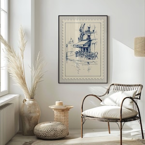 May include: A framed print depicting a blue and white sketch of a European cafe, designed to look like a postage stamp. The artwork is displayed in a bright room with a woven chair and decorative vase.