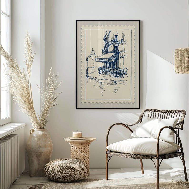 May include: A framed print depicting a blue and white sketch of a European cafe, designed to look like a postage stamp. The artwork is displayed in a bright room with a woven chair and decorative vase.