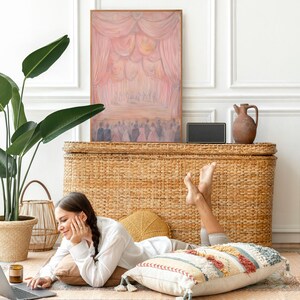May include: A woman is sitting on a rug in front of a wicker chest, using a laptop computer. She is wearing a white shirt and has her legs crossed. There is a plant in a pot to the left of the chest and a painting on the wall behind the chest.