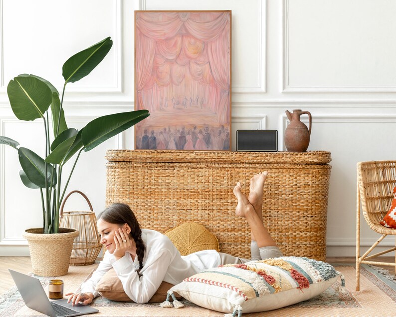 May include: A woman is sitting on a rug in front of a wicker chest, using a laptop computer. She is wearing a white shirt and has her legs crossed. There is a plant in a pot to the left of the chest and a painting on the wall behind the chest.